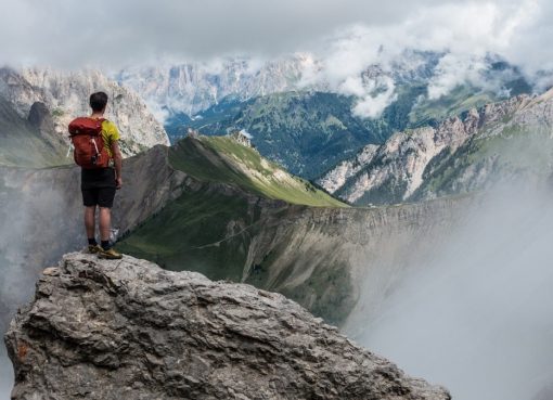 man with red backpack standing on cliff facing mountains under white sky during daytime